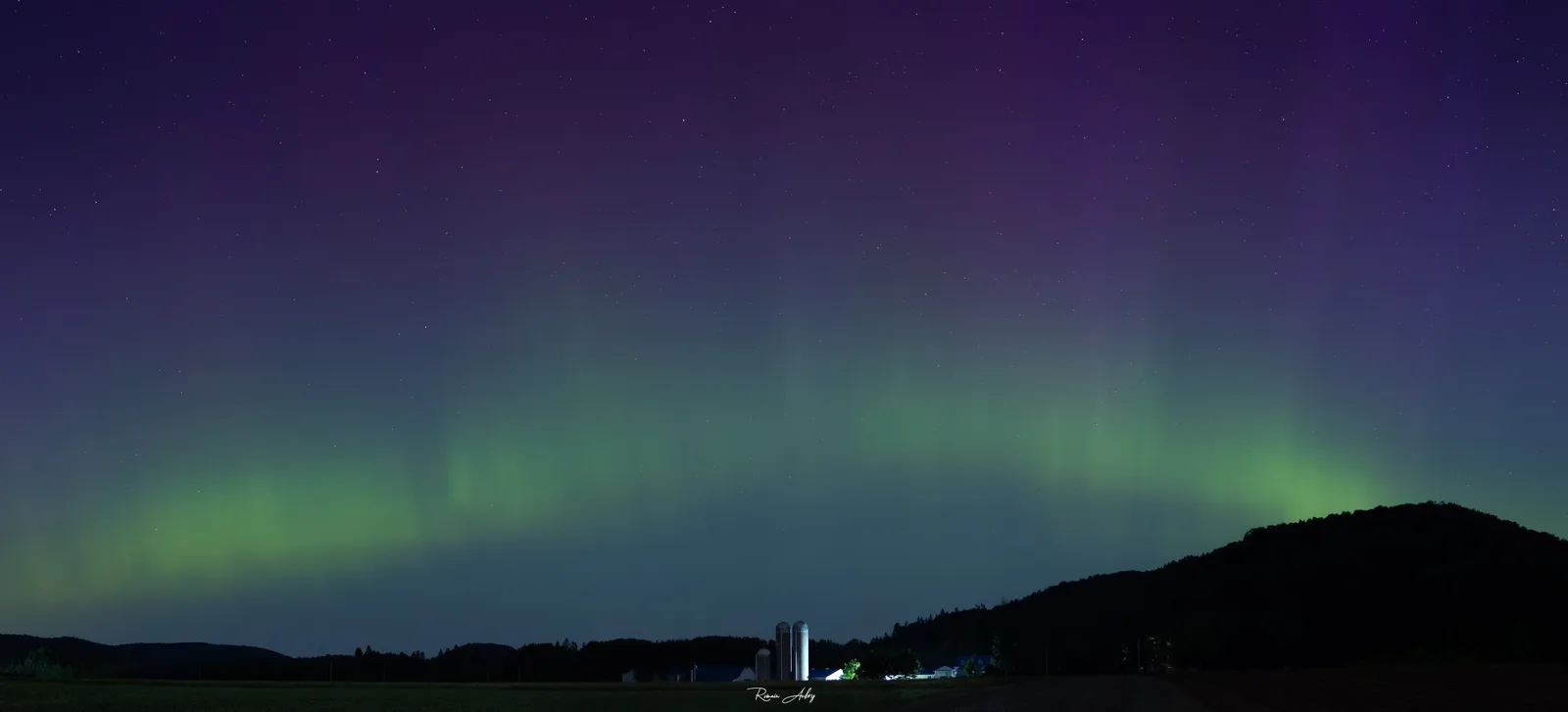 A green auroral arc low over a farm with silos, beneath a purple sky. A typical mid-latitude display seen from Quebec.